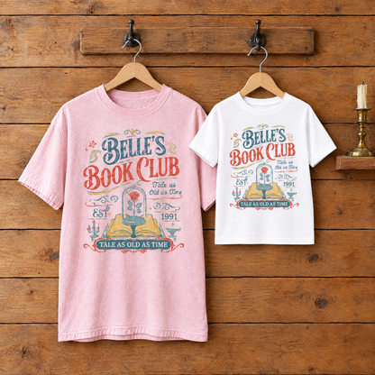 Adult Blossom Comfort Colors Belle's Book Club shirt and a White youth Belle's Book Club tee on wooden hangers