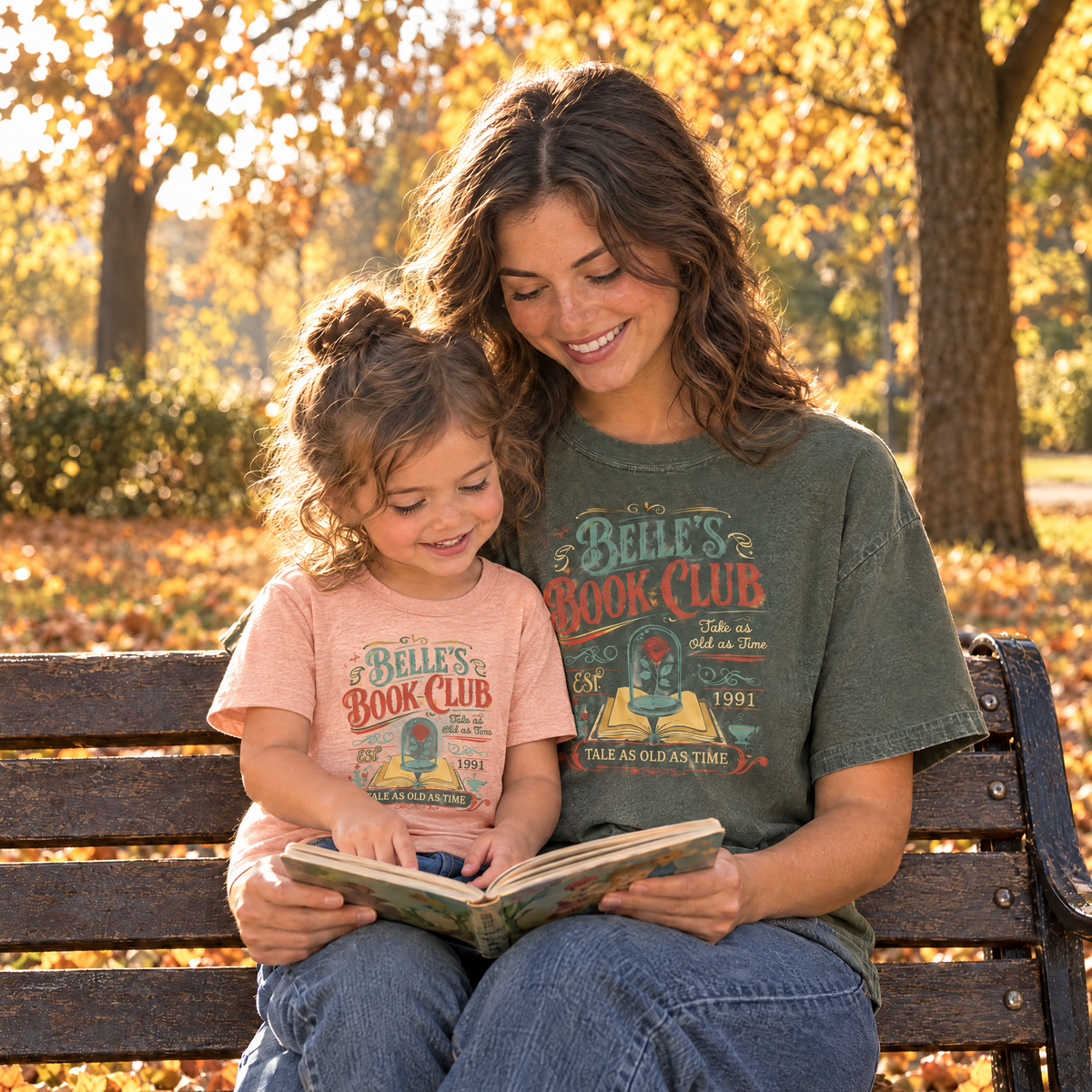 Mom in a Moss Comfort Colors Belle's Book Club shirt and toddler in a Heather Peach toddler Belle's Book Club tee reading on a park bench in autumn