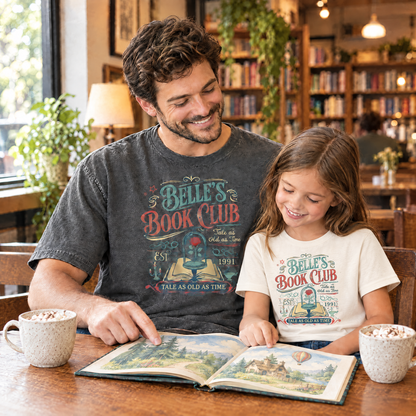 Dad in a Pepper Comfort Colors Belle's Book Club shirt and daughter in a Natural youth Belle's Book Club tee reading a picture book in a bookstore cafe
