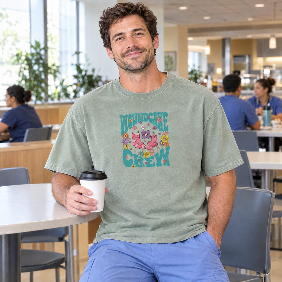 Male nurse wearing a Sage Comfort Colors Wound Care Crew shirt holding coffee in a hospital cafeteria