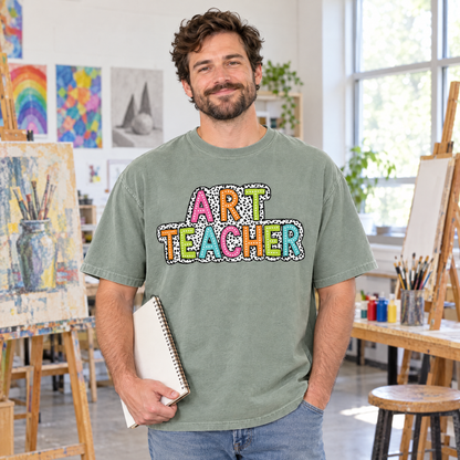Male art teacher wearing a Sage Comfort Colors Art Teacher shirt holding a sketchbook in a classroom