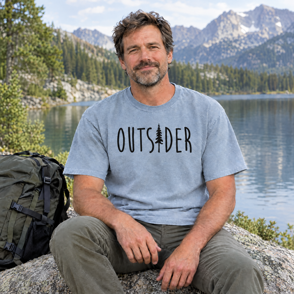 Outsider camping shirt with pine tree in Chambray Comfort Colors, man sitting on a rock by a mountain lake