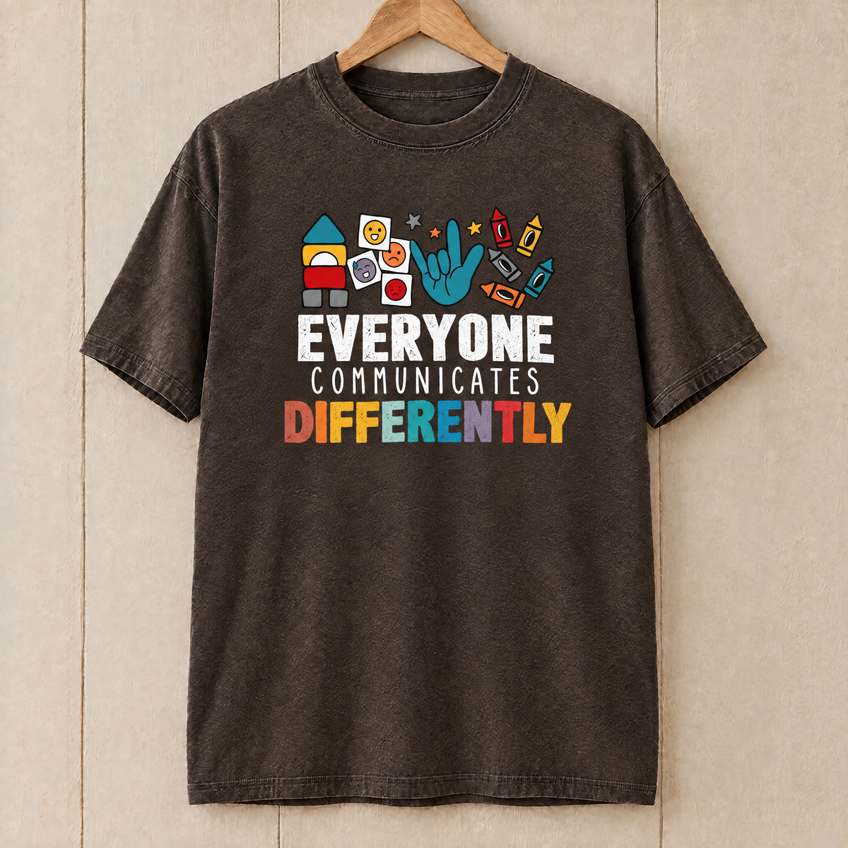 Everyone Communicates Differently autism acceptance shirt in Espresso Comfort Colors on a wooden hanger