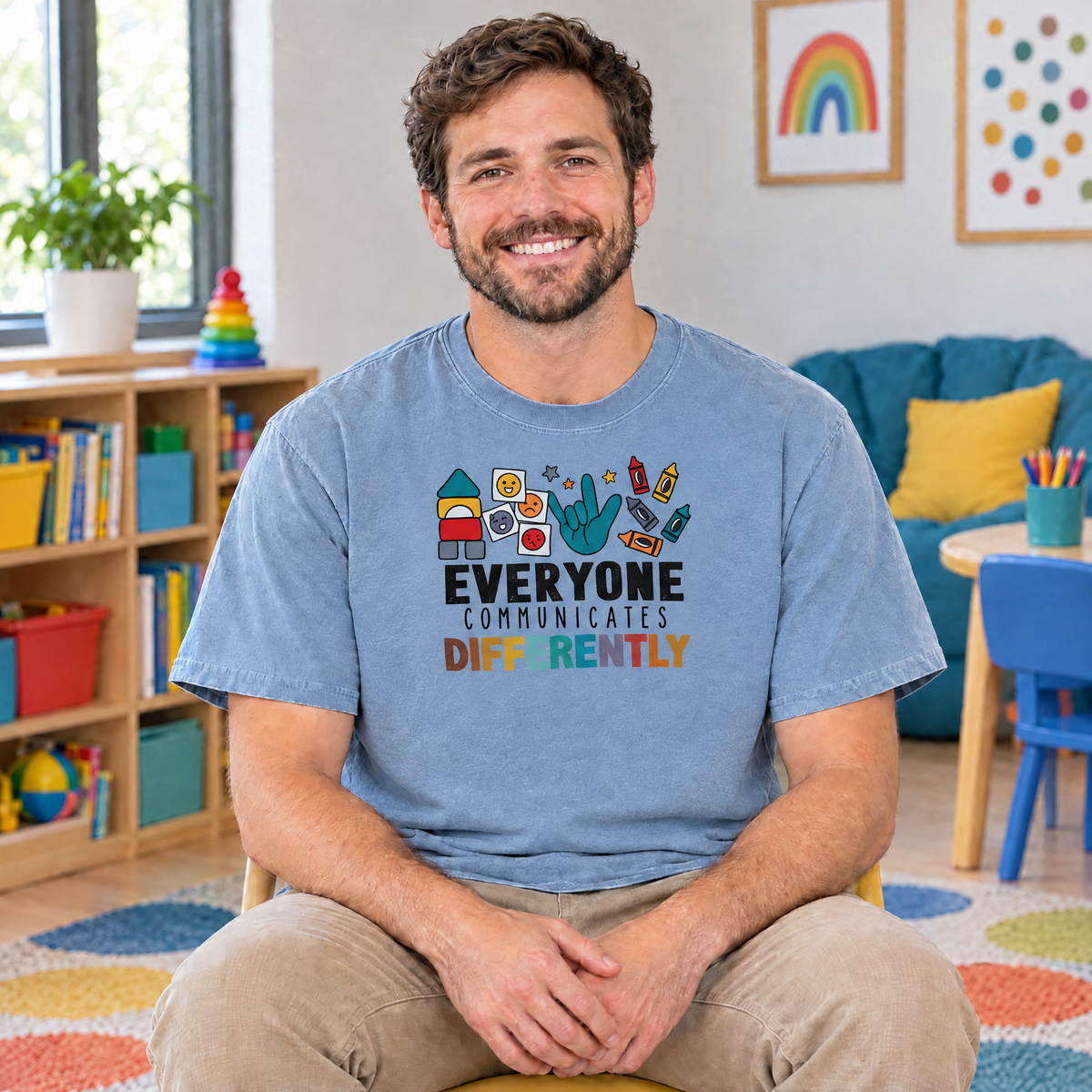 Everyone Communicates Differently autism acceptance shirt in Chambray Comfort Colors, speech therapist sitting in a colorful therapy room