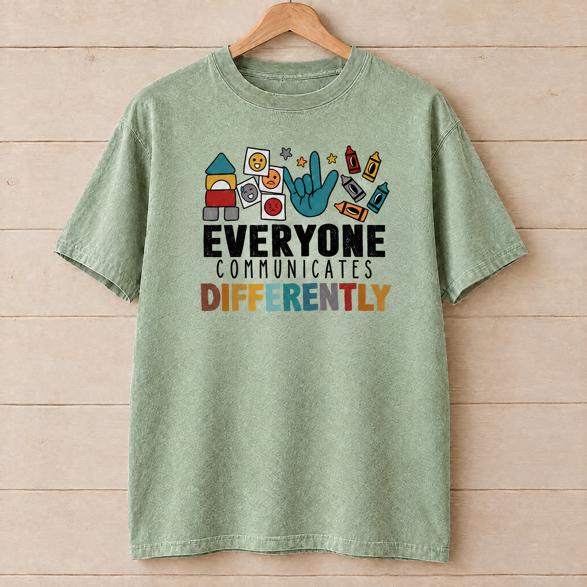 Everyone Communicates Differently autism acceptance shirt in Light Green Comfort Colors on a wooden hanger