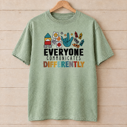 Everyone Communicates Differently autism acceptance shirt in Light Green Comfort Colors on a wooden hanger