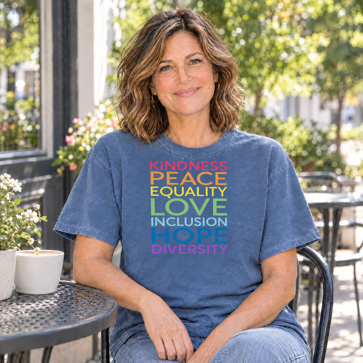 Kindness Peace Equality Love rainbow words shirt in Blue Jean Comfort Colors, woman sitting at an outdoor cafe table