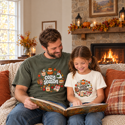 Dad in a Moss Comfort Colors Cozy Season shirt and daughter in a Natural youth Cozy Season tee reading by the fireplace