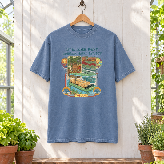 Blue Jean Get In Loser We're Learning About Lettuce shirt on a wooden hanger in a sunny greenhouse