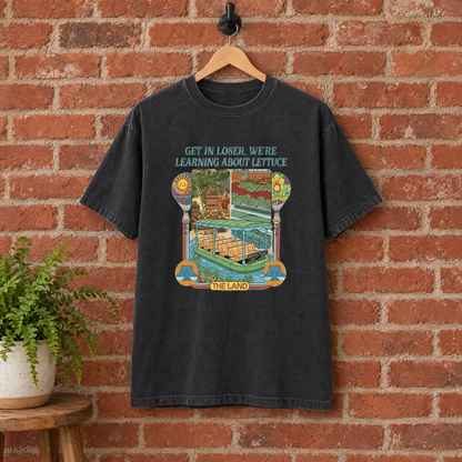Black Learning About Lettuce shirt on a hanger against a red brick wall