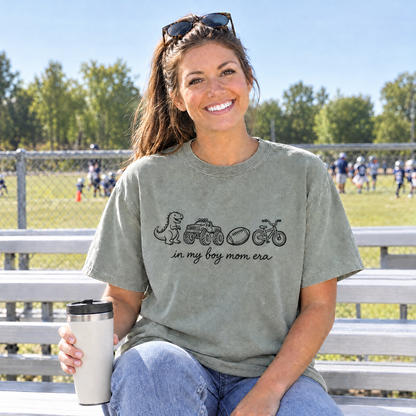 Football mom in a Sage In My Boy Mom Era shirt sitting on the bleachers at a youth game