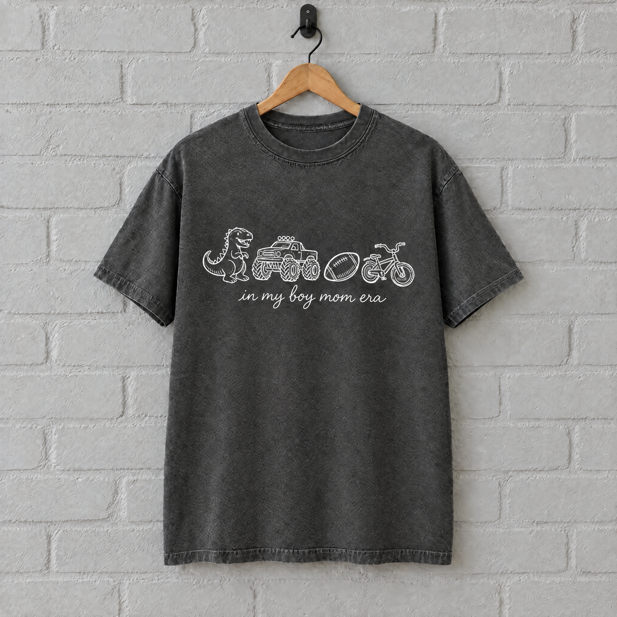 Pepper grey boy mom shirt with white print on a hanger against a painted brick wall