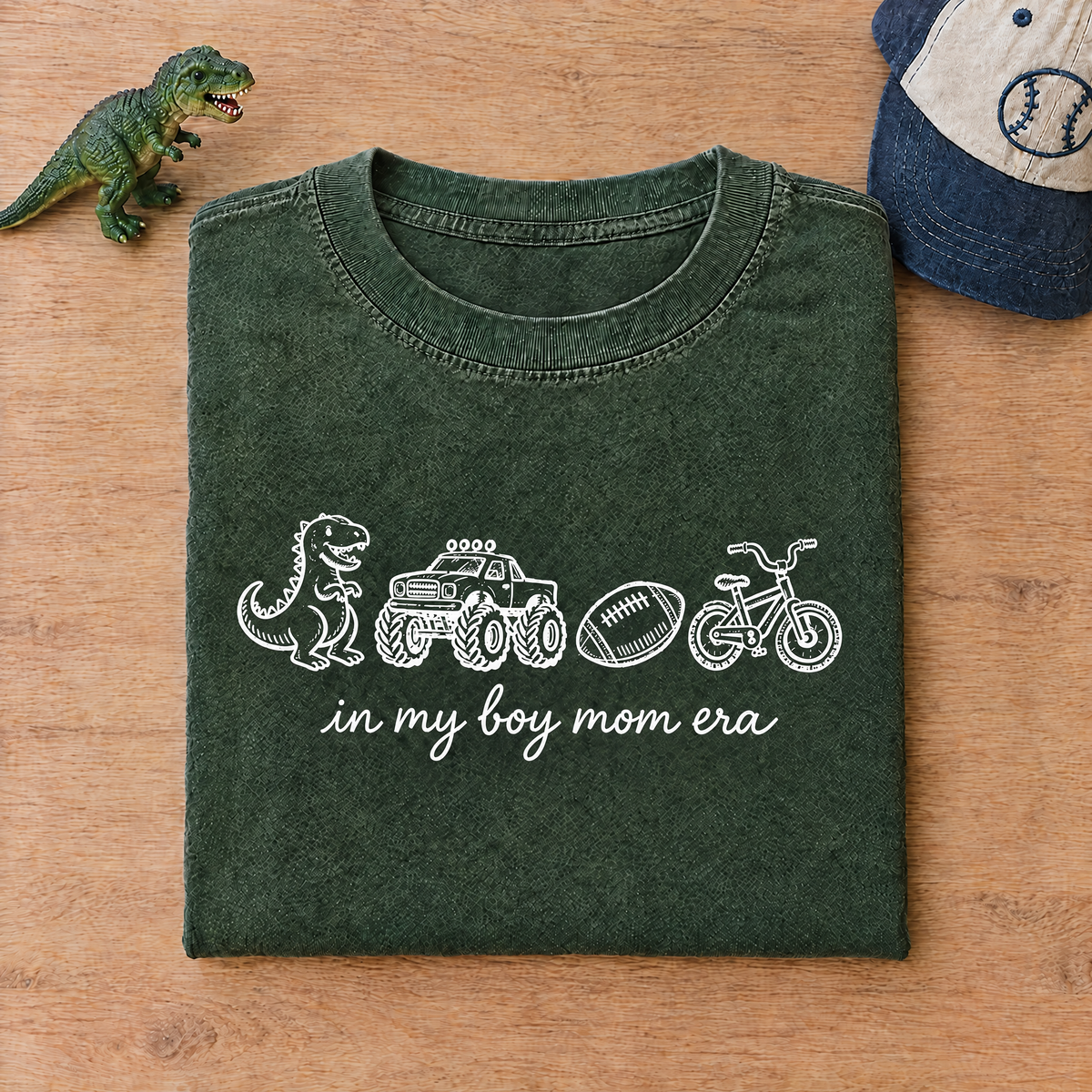 Moss green boy mom shirt folded next to a toy dinosaur and a kids cap