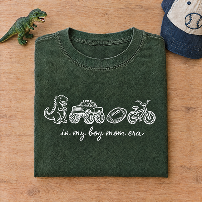 Moss green boy mom shirt folded next to a toy dinosaur and a kids cap