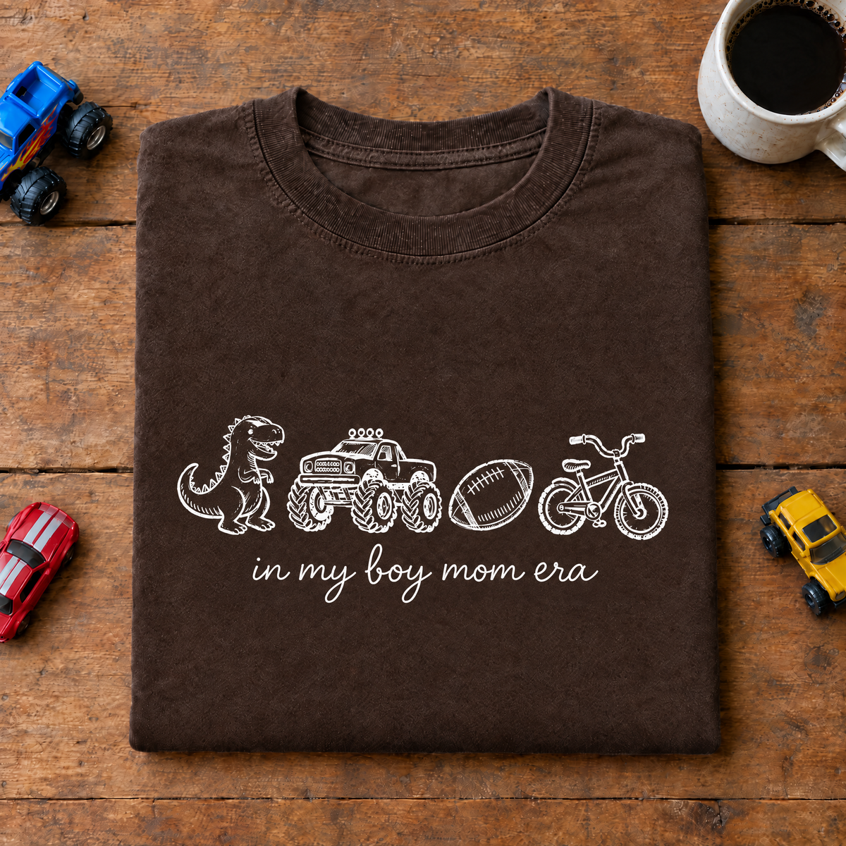 Espresso brown In My Boy Mom Era shirt folded on a wooden table with toy cars and coffee