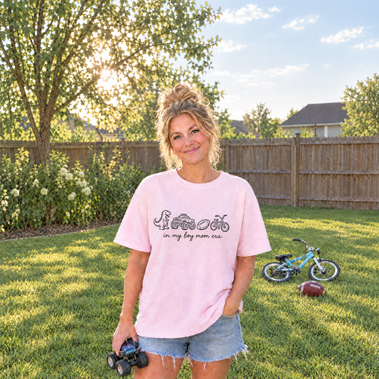 Mom in a Blossom pink In My Boy Mom Era shirt with dinosaur, monster truck, football and bike in a sunny backyard