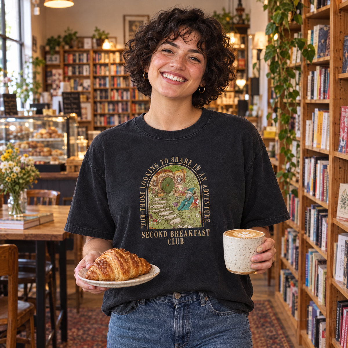 Woman wearing Black Comfort Colors Second Breakfast Club shirt in a bookstore cafe with a croissant