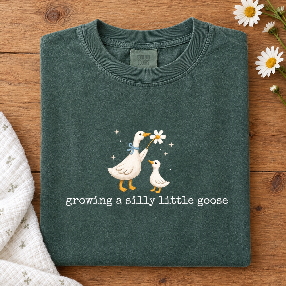 Silly goose pregnancy tee in Blue Spruce, folded with a baby blanket and daisies