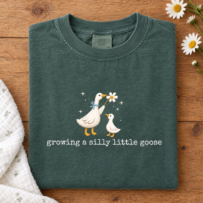 Silly goose pregnancy tee in Blue Spruce, folded with a baby blanket and daisies