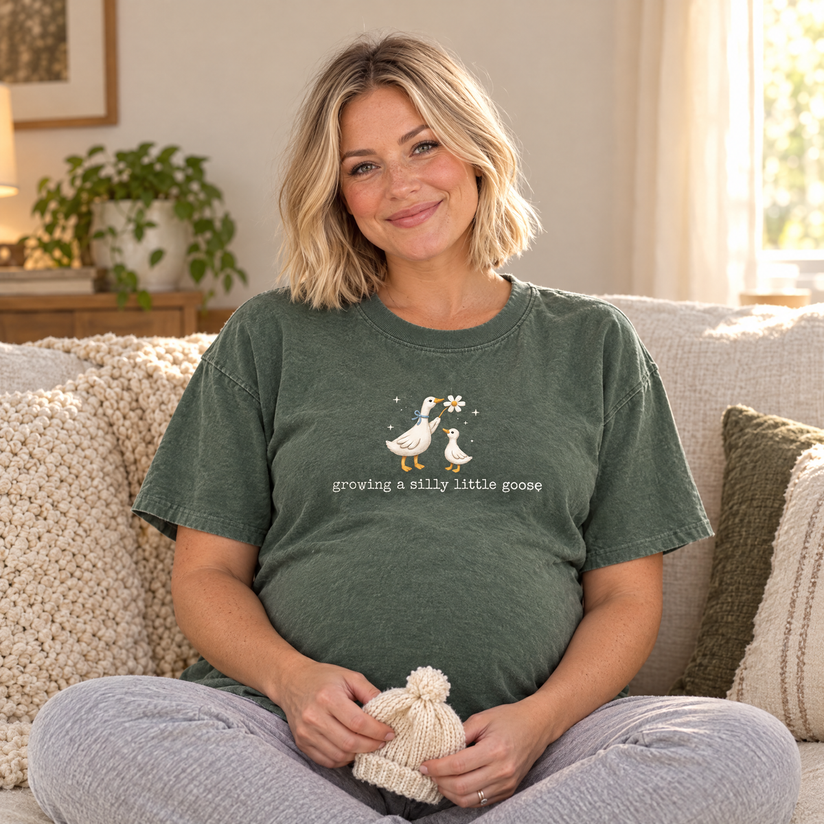 Pregnant woman wearing Moss Comfort Colors silly goose shirt on the sofa holding a knitted baby hat